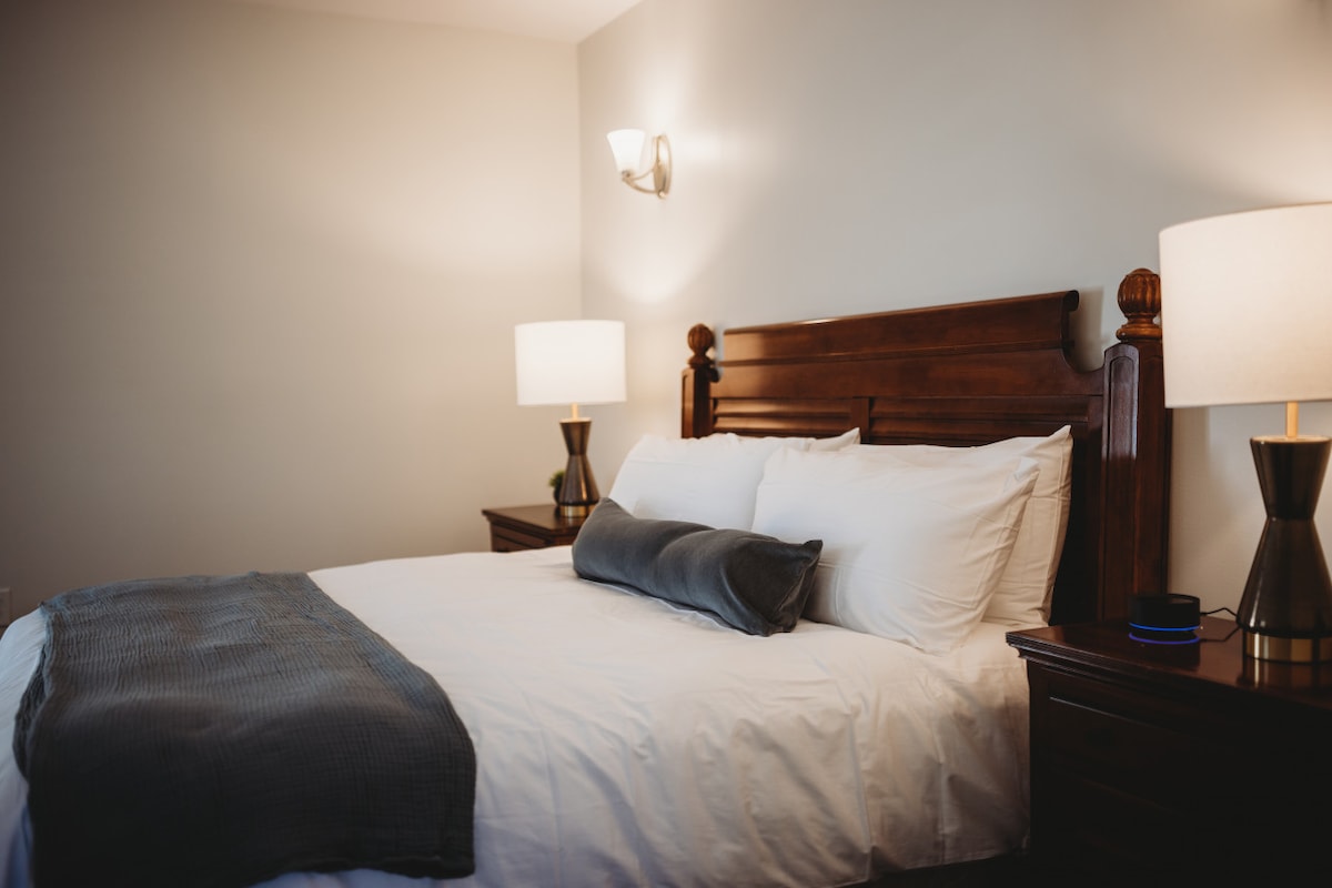 A spacious guest room is designed with a wooden bed frame and crisp white linens. Soft gray pillows accentuate the bed. Nightstands with modern lamps provide gentle lighting, enhancing the serene atmosphere of the space.