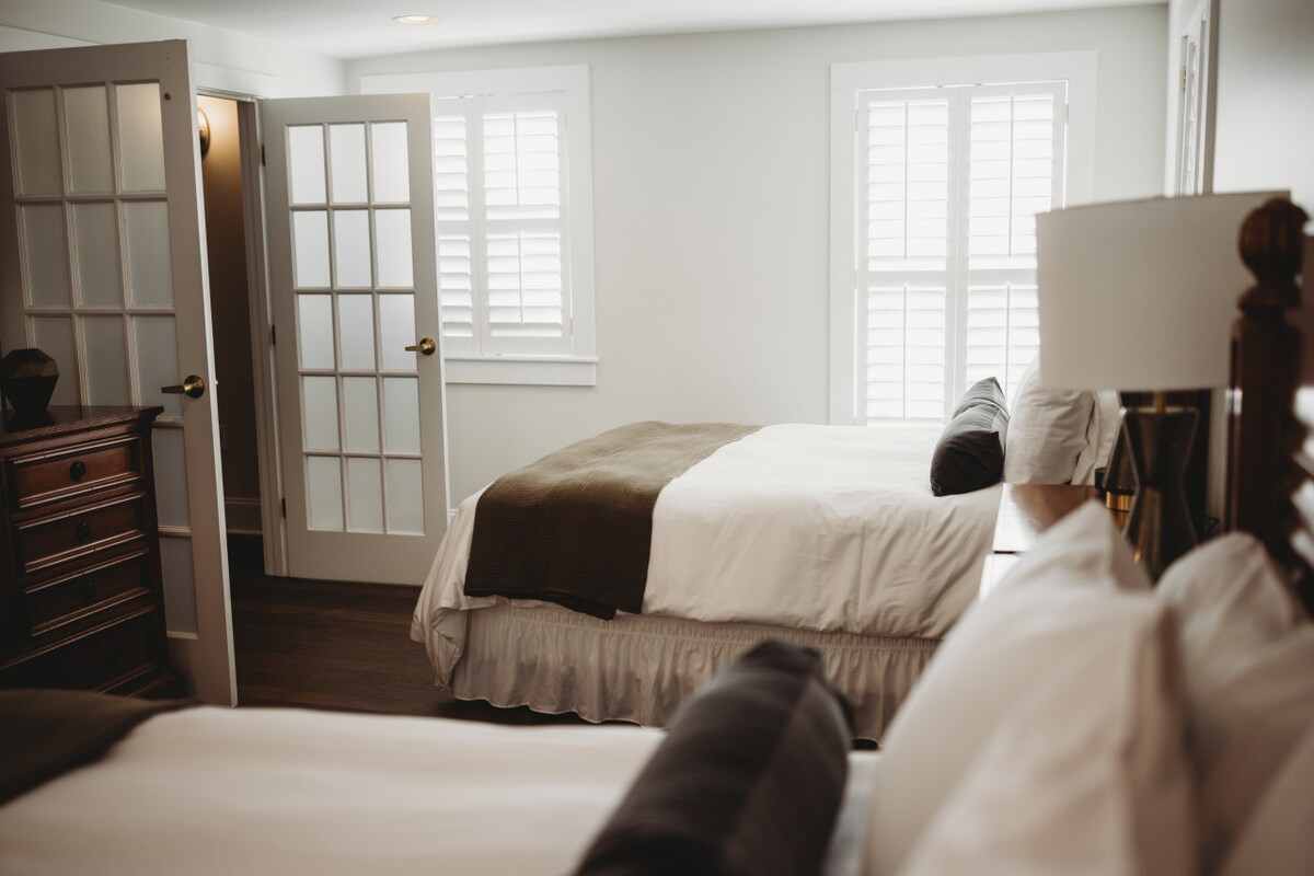 A spacious suite is shown, featuring two queen beds with white bedding and dark accent pillows. Covered French doors lead to another room, while light streams through the partially opened window, highlighting the warm wooden furnishings and neutral tones of the decor.