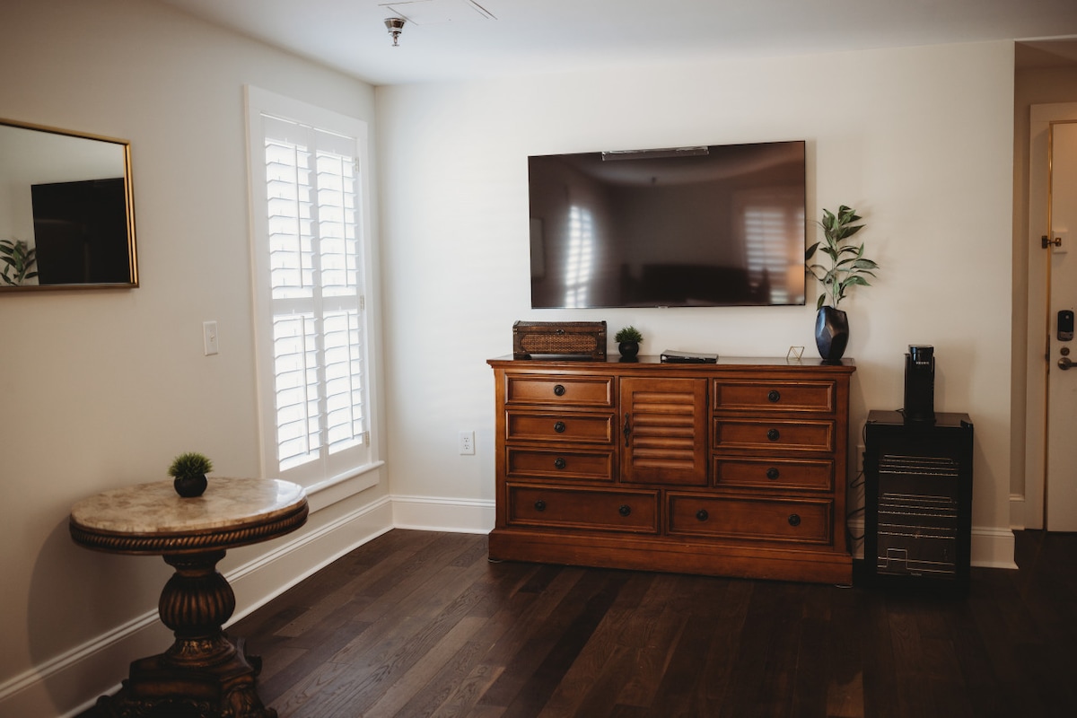 A well-lit living space features a wooden dresser with multiple drawers beneath a flat-screen television. A small round side table is positioned nearby, adorned with a decorative plant. Light-filtering shutters are present on the window, enhancing the room's brightness.
