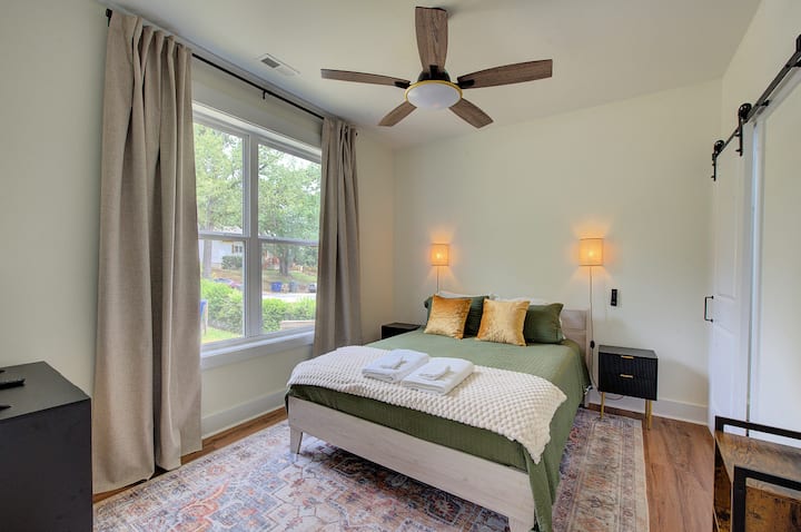 Comodidad moderna: dormitorio elegante con ropa de cama de color verde tierra, almohadas de lujo con detalles dorados y grandes ventanales para que entre la luz natural y el aire fresco.