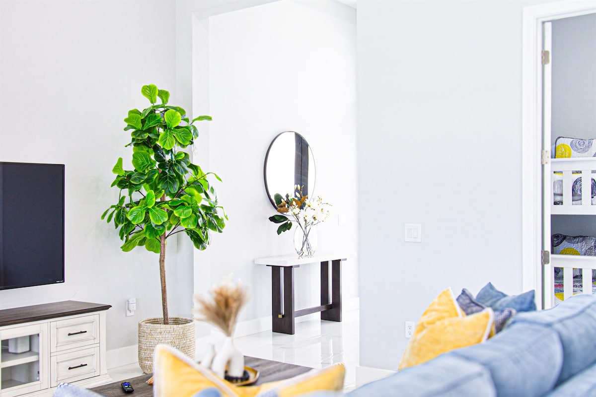 A spacious living area is highlighted by a flat-screen television mounted on the wall. A decorative plant in a woven basket adds a touch of greenery. Light-colored walls and elegant flooring create an open atmosphere, with a mirror and console table visible in the background.