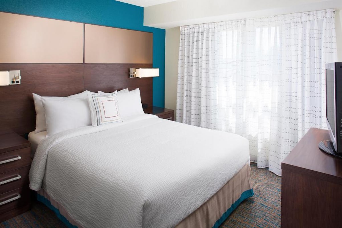 A well-appointed bedroom features a spacious bed with crisp white bedding and decorative pillows. A blue accent wall complements the warm wood furnishings. Sheer curtains allow light to filter in from a large window, enhancing the room's bright and airy feel.