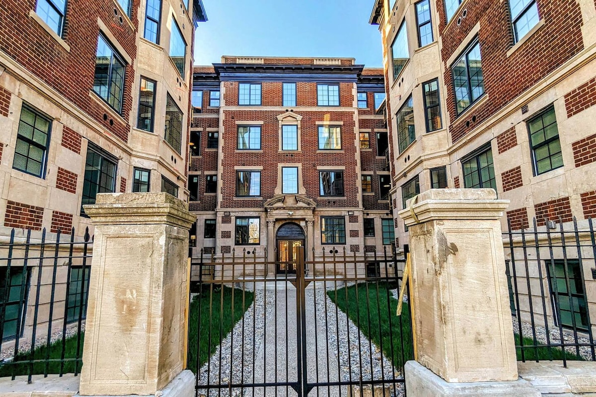A historic brick building showcases elegant architectural details, including large windows and a prominent entrance. Lush green grass surrounds a central courtyard, framed by decorative stone gate pillars. Soft morning light highlights the structure's classic charm.