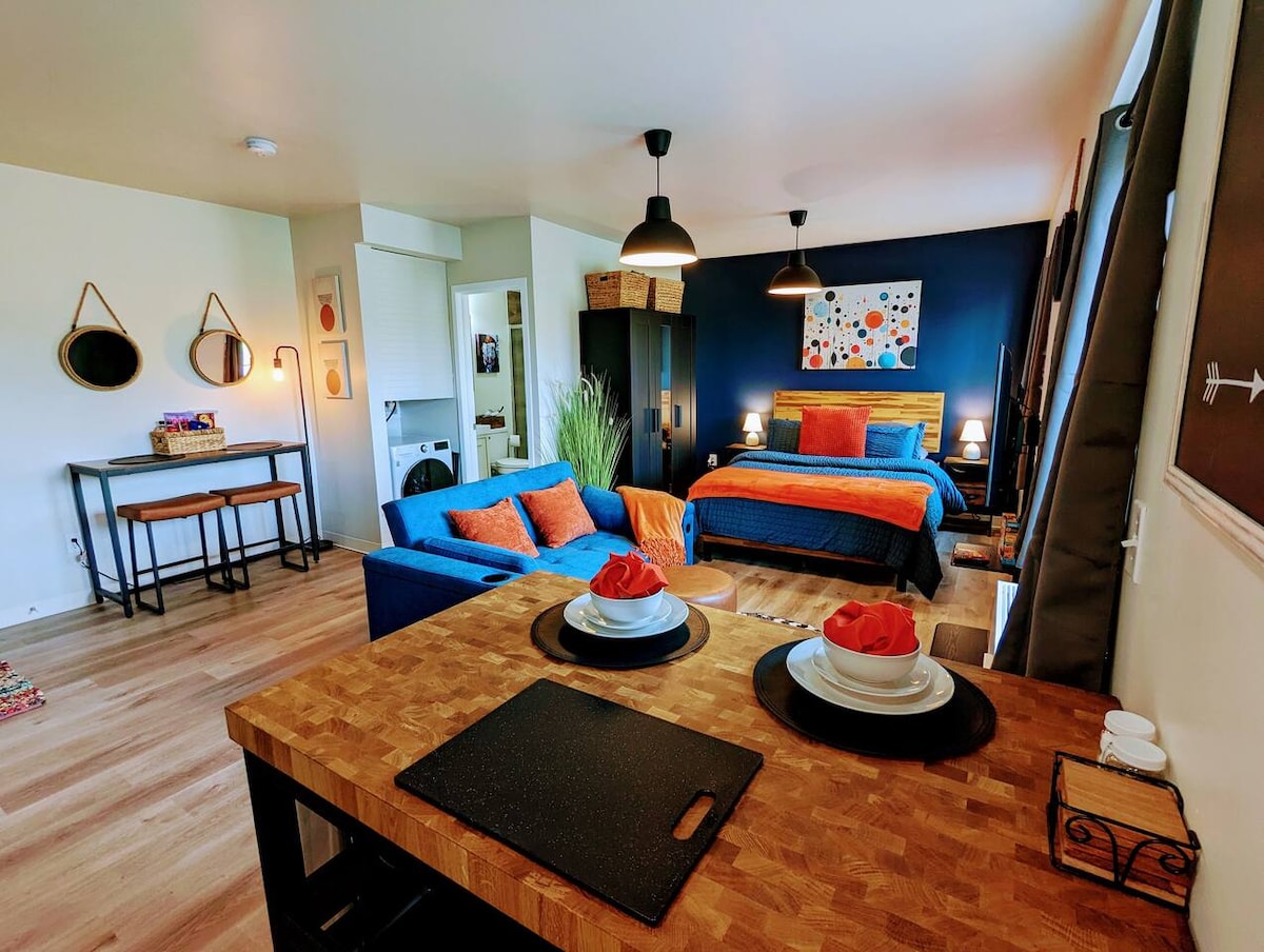 A bright studio space features a queen bed with a colorful accent wall and a blue sofa bed. A wooden dining table is set with plates and rose decorations. Light fixtures create a warm ambiance, while mirrors and artwork enhance the decor.