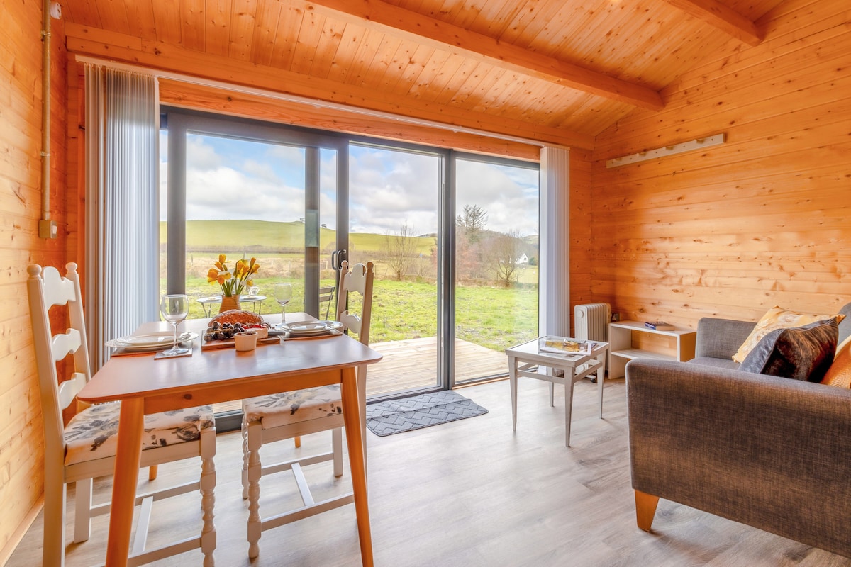 An open plan living space showcases a dining table set for two, featuring a floral centerpiece. Large sliding doors provide a view of the countryside and open onto an outdoor deck. Warm wooden paneling and natural light create a welcoming atmosphere in the cozy cabin.