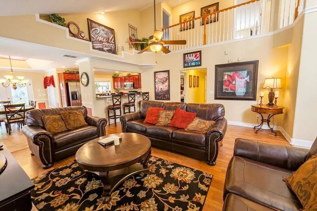 A spacious living area features two brown leather sofas situated around a circular wooden coffee table. A large area rug with intricate patterns covers the floor. Ceiling fans enhance the open concept, with warm light illuminating colorful artwork on the walls.