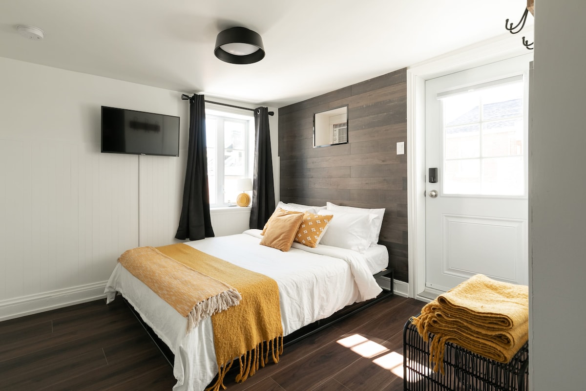 A warm sleeping area features a spacious bed dressed in white linens, complemented by a mustard throw and decorative pillows. Natural light enters through a window dressed with dark curtains, while a mounted television is positioned on the wall, next to a door leading outside.
