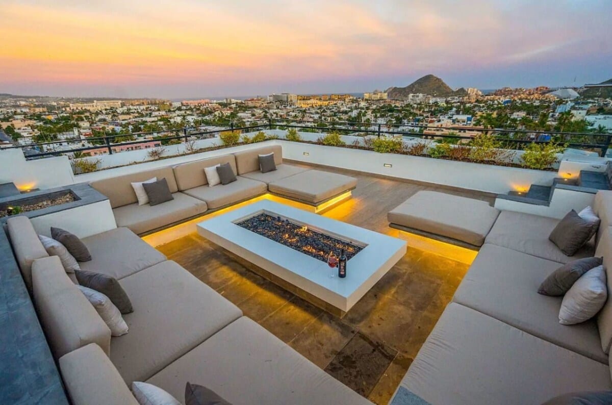 An exclusive rooftop terrace features a spacious seating area arranged around a centrally located fire pit. Cushioned benches invite relaxation, while the surrounding views showcase a vibrant sunset over the city landscape, creating a serene atmosphere for gatherings.