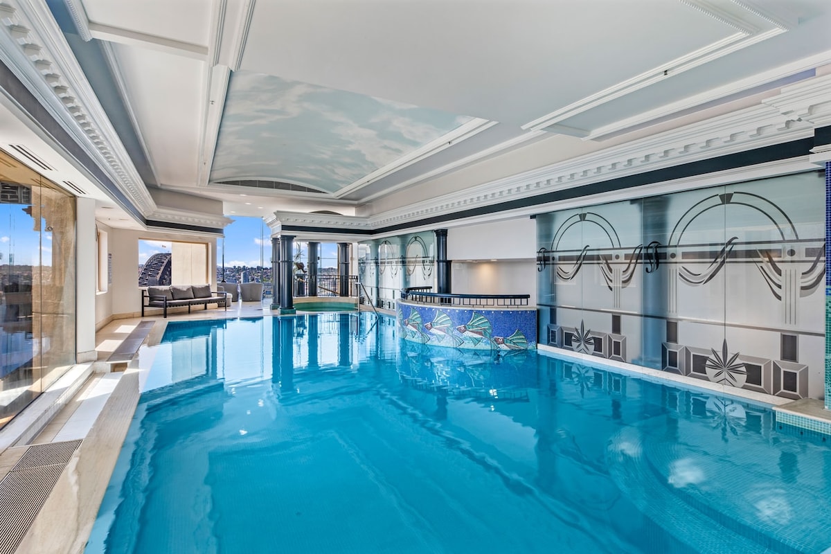 A spacious indoor pool area features clear blue water reflecting the ceiling’s cloud mural. The elegant design includes decorative tile work and large windows that provide artificial and natural light, creating a serene atmosphere. Comfortable seating is arranged alongside the pool.