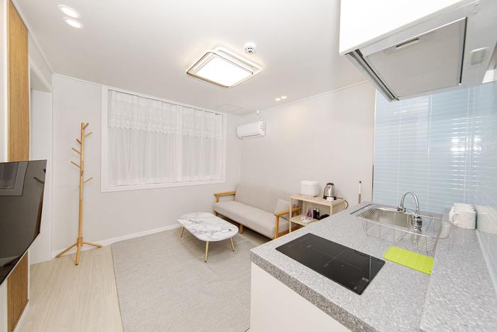 A bright living area is displayed, featuring a light-colored sofa resting against the wall. A small, round coffee table is positioned in front, on a soft area rug. The kitchenette is visible in the background, equipped with modern appliances and a simple countertop.