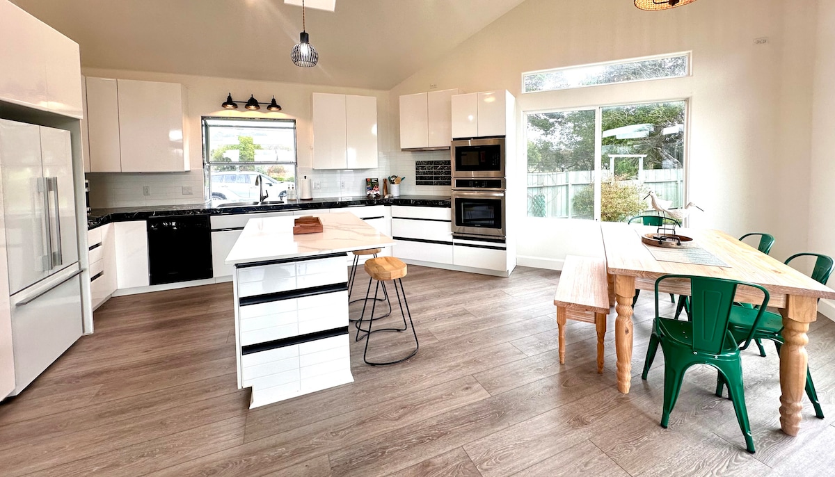 A bright and modern kitchen is presented with sleek white cabinetry and stainless steel appliances. A spacious island with bar seating complements the dining area featuring a wooden table and green chairs. Large windows allow natural light to fill the space, offering views of the outdoors.