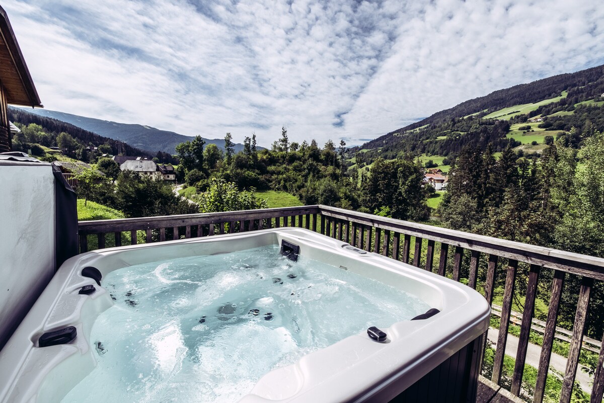 A hot tub is situated on a balcony with a wooden railing, providing expansive views of rolling green hills and distant mountains. The sky is filled with soft clouds, enhancing the serenity of the rural surroundings.