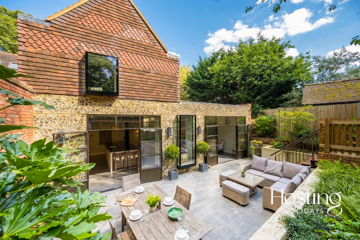 The outdoor space features a paved patio furnished with a large sectional sofa, dining table, and chairs. Sliding glass doors open to the interior, allowing for easy access. Lush greenery and trees provide a natural backdrop, enhancing the relaxing atmosphere of the area.