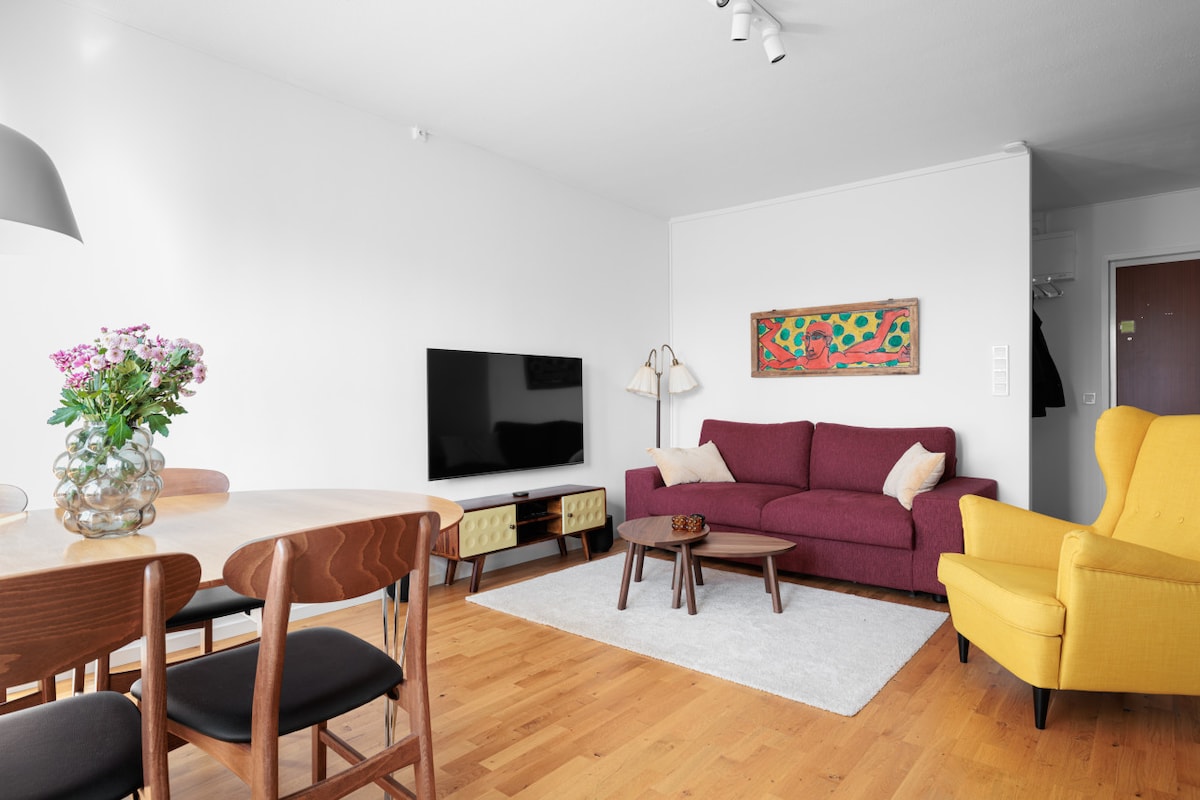 The living area features a comfortable maroon sofa, a modern yellow armchair, and a wooden dining table set for four. A large television is positioned on a low TV stand, while a colorful artwork adds a touch of brightness to the neutral walls. Natural light fills the room.
