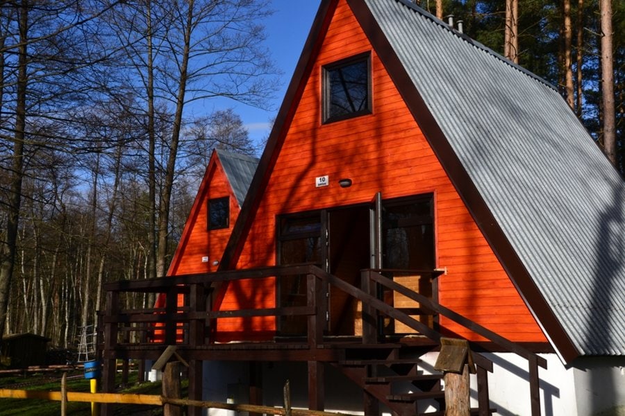 A pair of triangular-roofed cabins with vibrant orange exteriors is set amidst a wooded area. Each cabin features two large windows on the upper level, allowing natural light to illuminate the interior. A wooden deck with railings provides access to the entrance of one cabin.