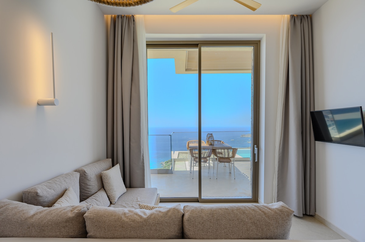 A spacious living area is featured, complete with a comfortable sectional sofa facing large glass doors showcasing panoramic sea views. Light drapes frame the entrance to a private terrace, where outdoor dining furniture is visible against the backdrop of the tranquil blue sea.