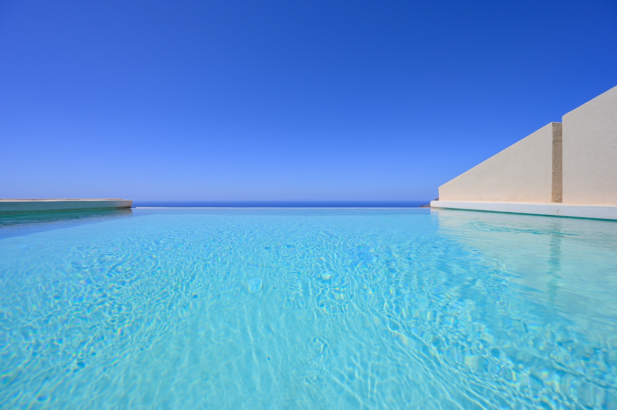 An infinity pool with clear turquoise water provides stunning views of the Cretan Sea against a backdrop of a cloudless blue sky. The pool's edges appear to blend seamlessly with the horizon, creating a tranquil atmosphere.