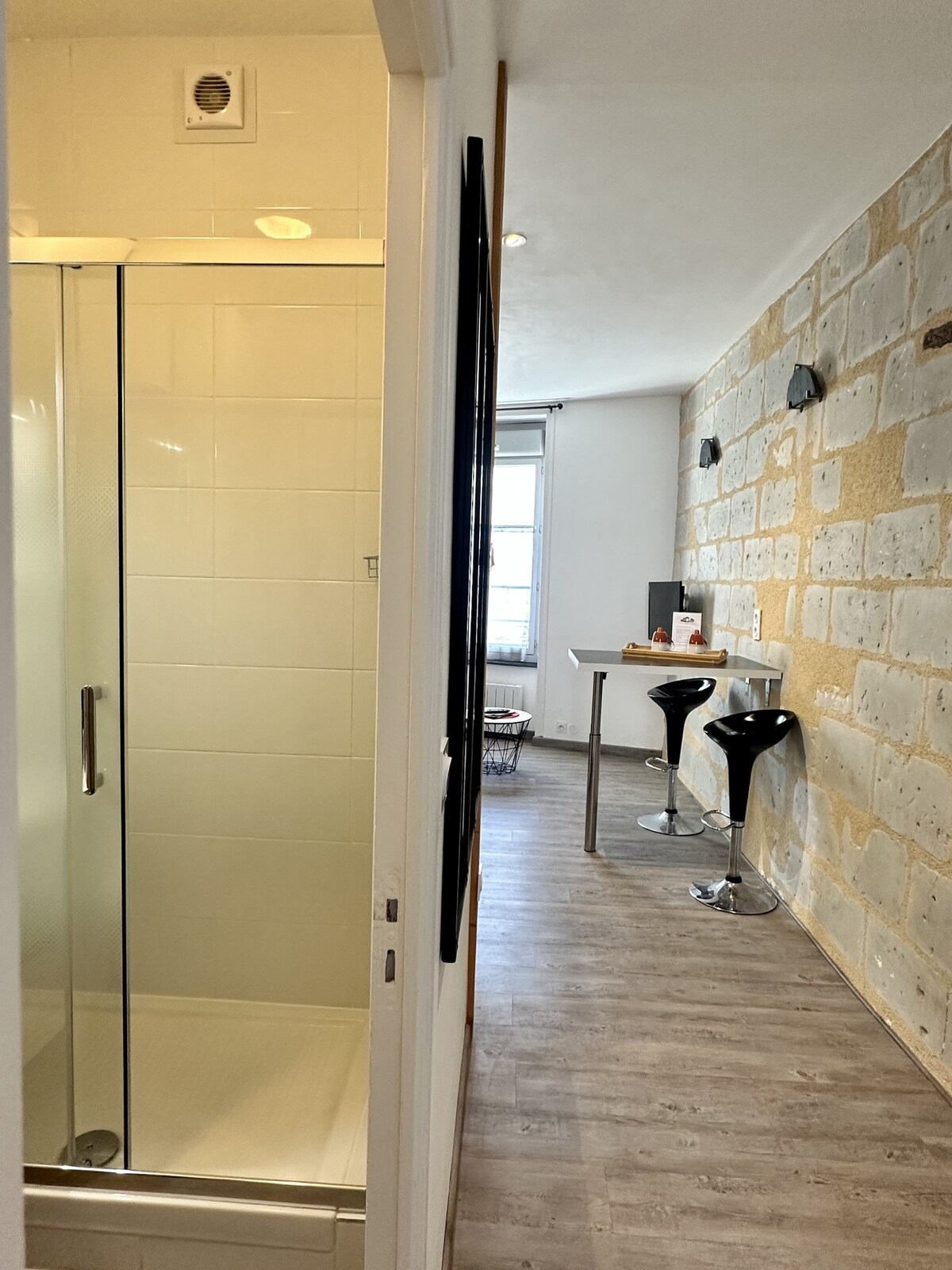 A view of the bathroom area includes a modern glass shower enclosure with white tiles. Beyond, a dining space is visible, featuring a compact table with two black bar stools against a charming stone wall, which adds texture to the interior.