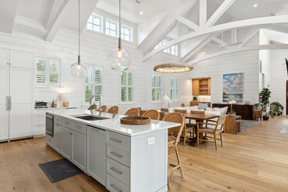 A spacious kitchen and dining area is highlighted by vaulted ceilings and large windows allowing natural light. The kitchen features a central island with seating, while a dining table accommodates several chairs. Light wood flooring complements the bright, airy decor.