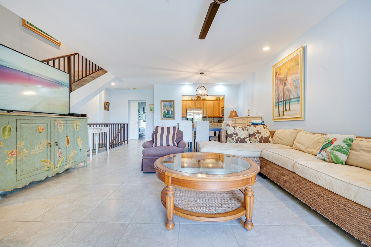 An open living area features a large sectional sofa with tropical-themed cushions, a round wicker coffee table, and a modern television on a decorative console. Warm lighting and beach-themed artwork complement the spacious environment, enhancing comfort and relaxation.