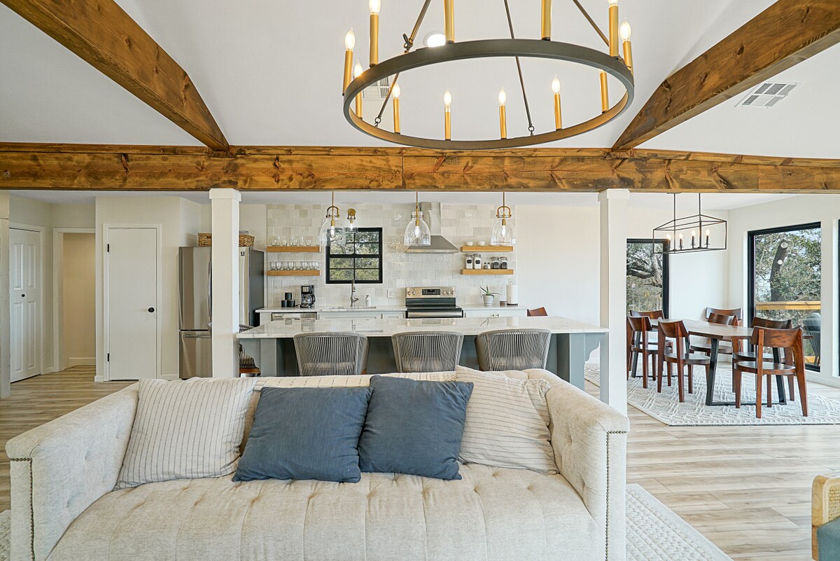 An open-concept living area showcases wooden beams overhead and modern furnishings. A light-colored couch with decorative pillows faces the kitchen, featuring a large island with bar stools. Dining options are visible in the background, with a wooden table set for meals, and large windows framing scenic views.