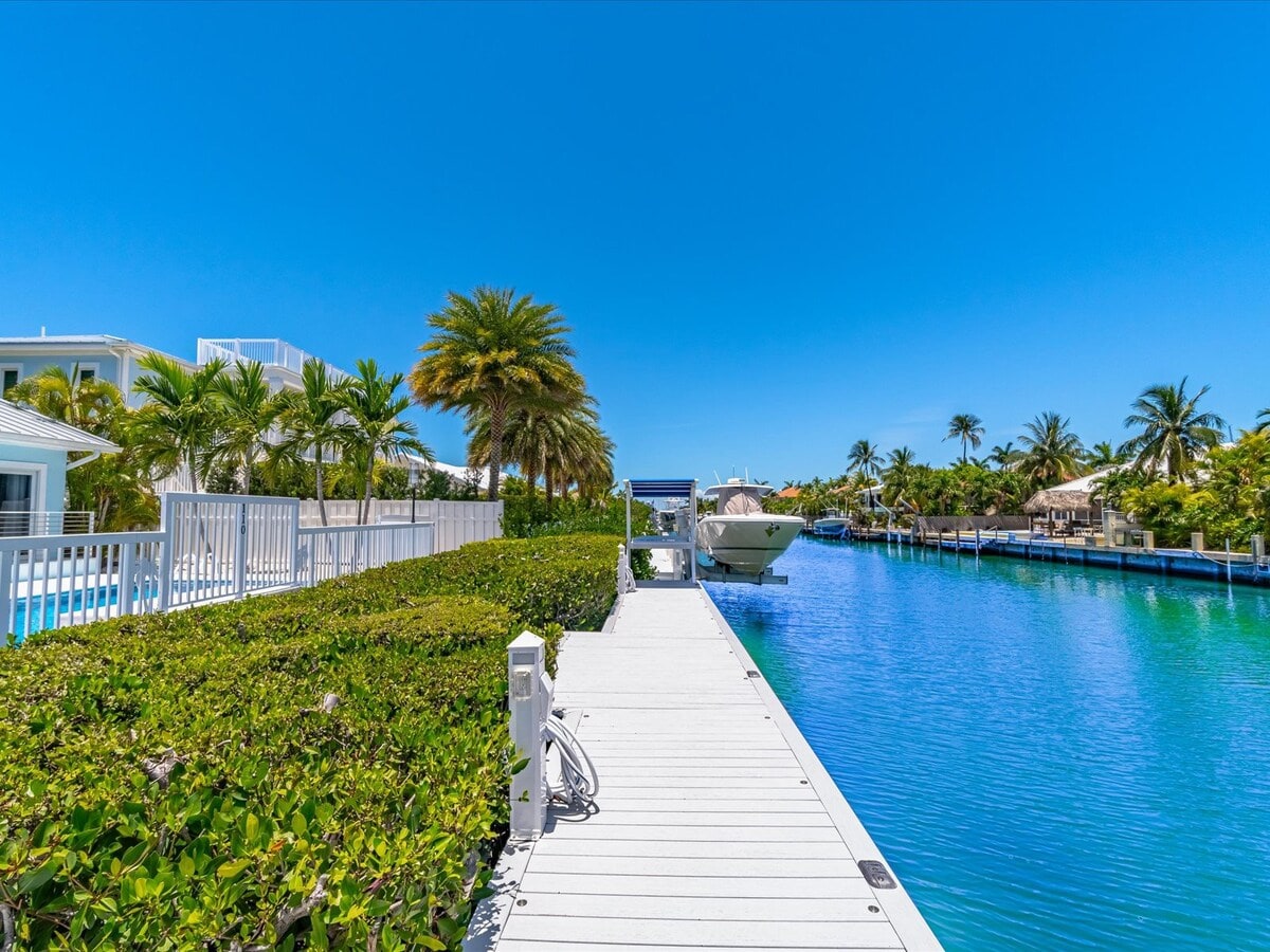 A private dock extends over tranquil waters, bordered by lush greenery and palm trees. The bright blue sky reflects on the water's surface, while a boat is visible at the end of the dock, inviting guests to explore the surrounding waterways.