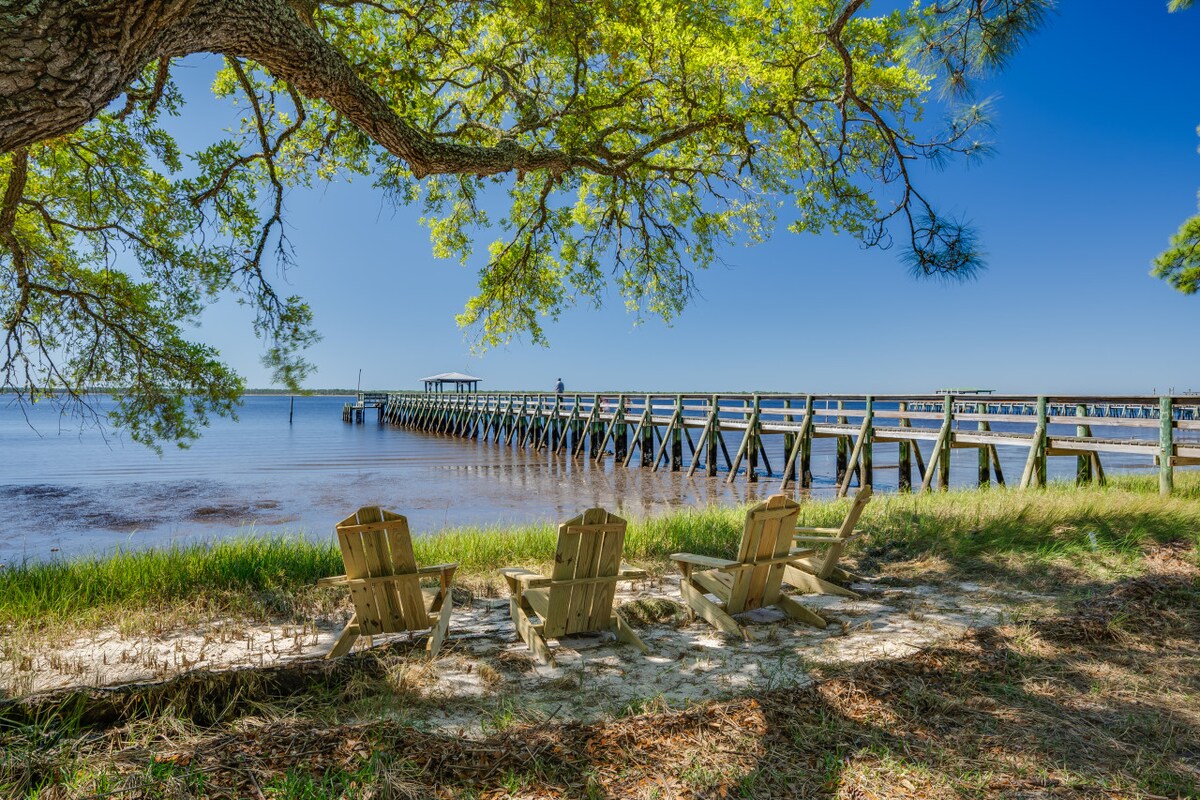 Three outdoor chairs are positioned on the grassy shore, providing a view of the dock extending into the calm waters. Lush greenery surrounds the area, and a large tree offers shade on a clear, sunny day.