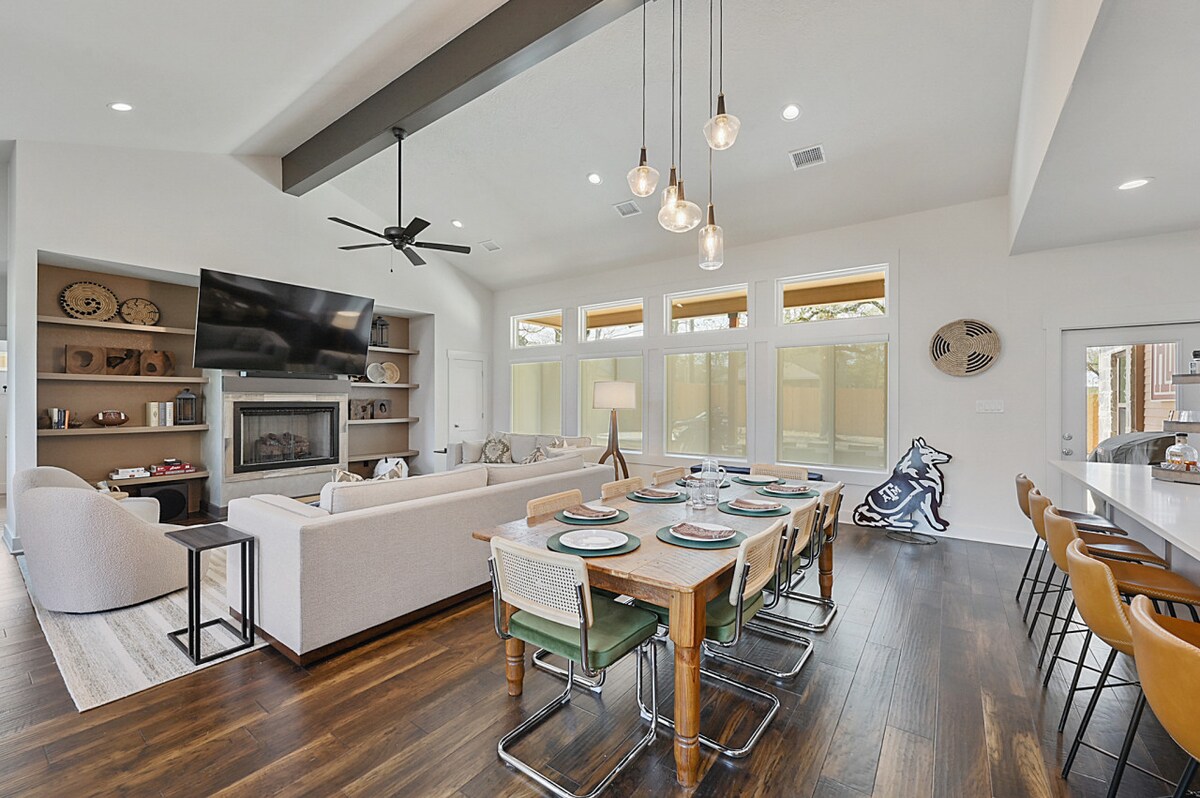 A spacious living area showcases a large sectional sofa and a mounted TV above a fireplace, complemented by natural light from numerous windows. A dining table with colorful seating is set for meals, while modern pendant lights add a contemporary touch to the open design.