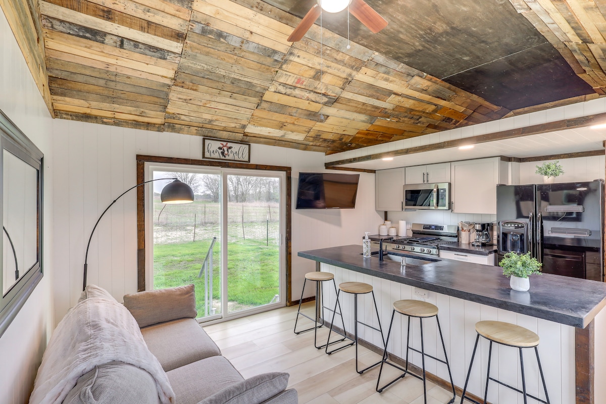 An open living area is showcased with natural light filtering through large windows. A modern kitchen with stainless steel appliances is complemented by a dining bar featuring four stools. A sofa rests nearby, creating a comfortable gathering space.