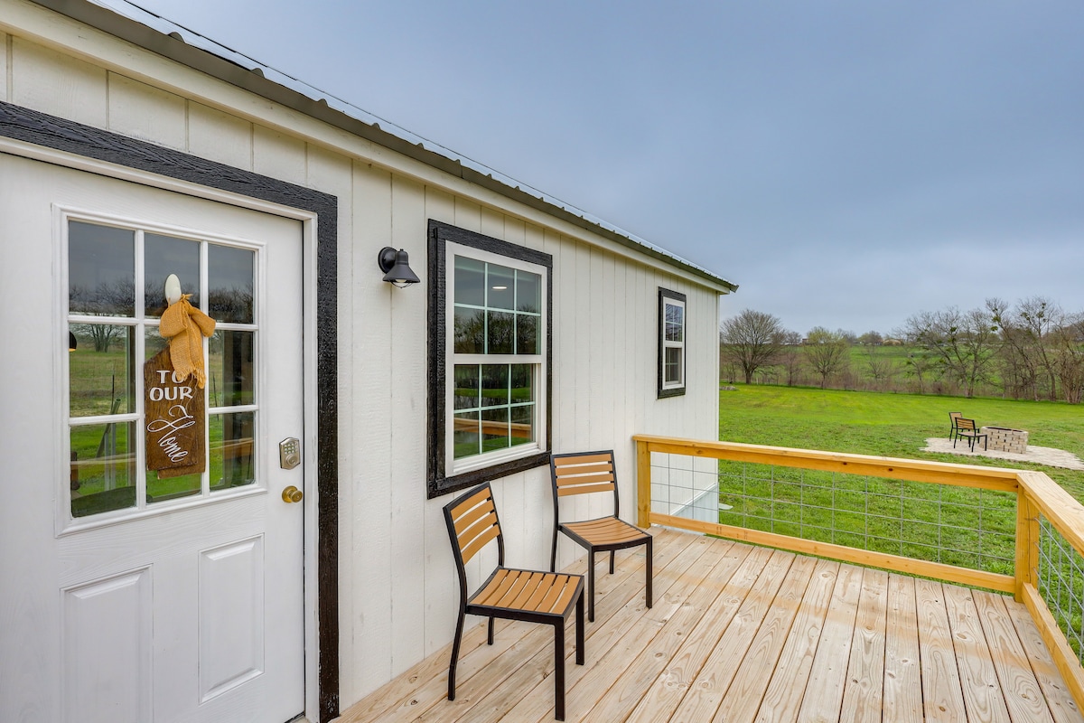 A wooden deck is adorned with two chairs, offering views of the surrounding green landscape. The entrance is marked by a white door with a decorative welcome sign. Large windows provide natural light and a connection to the outdoors.
