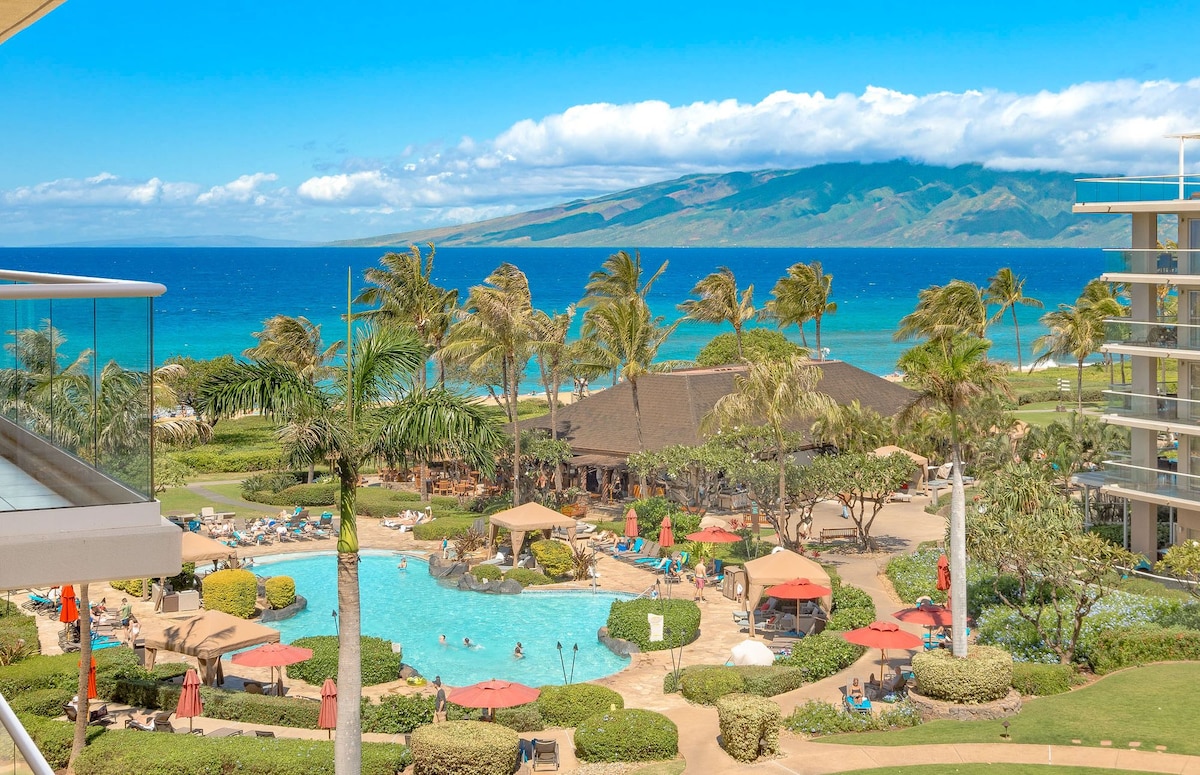 The image captures a view from the 5th-floor balcony, overlooking a vibrant pool area surrounded by lush greenery. Palm trees sway in the breeze, with the turquoise Pacific Ocean and the island of Molokai visible in the distance under a clear blue sky.