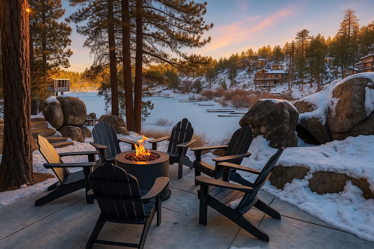An outdoor fire pit area is surrounded by multiple black adirondack chairs, positioned on a smooth concrete surface. Nearby snowy boulders and tall pine trees create a natural backdrop, while the serene lake view is visible, illuminated by the warm light of a sunset.