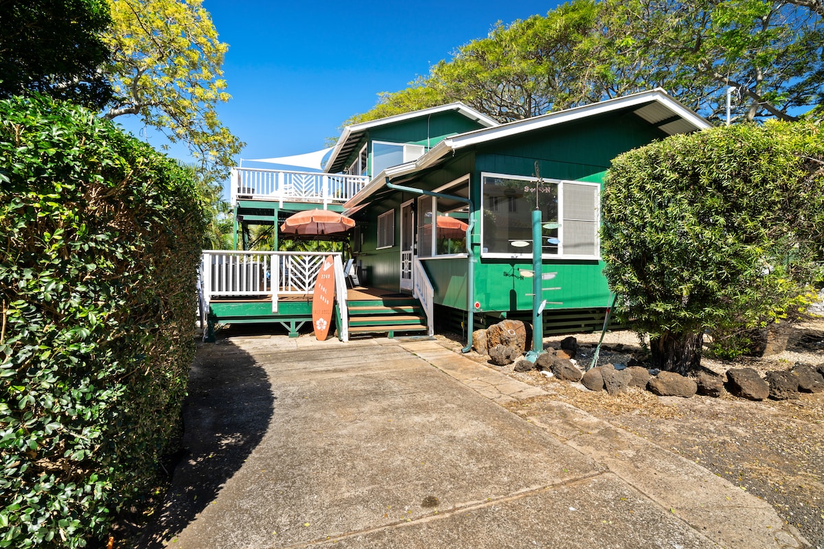 A lush green home with a welcoming exterior is highlighted by a spacious front porch and multiple outdoor seating areas. The driveway leads to the entrance, bordered by well-maintained shrubs and trees. Bright blue skies frame the scene, enhancing the home's tropical charm.
