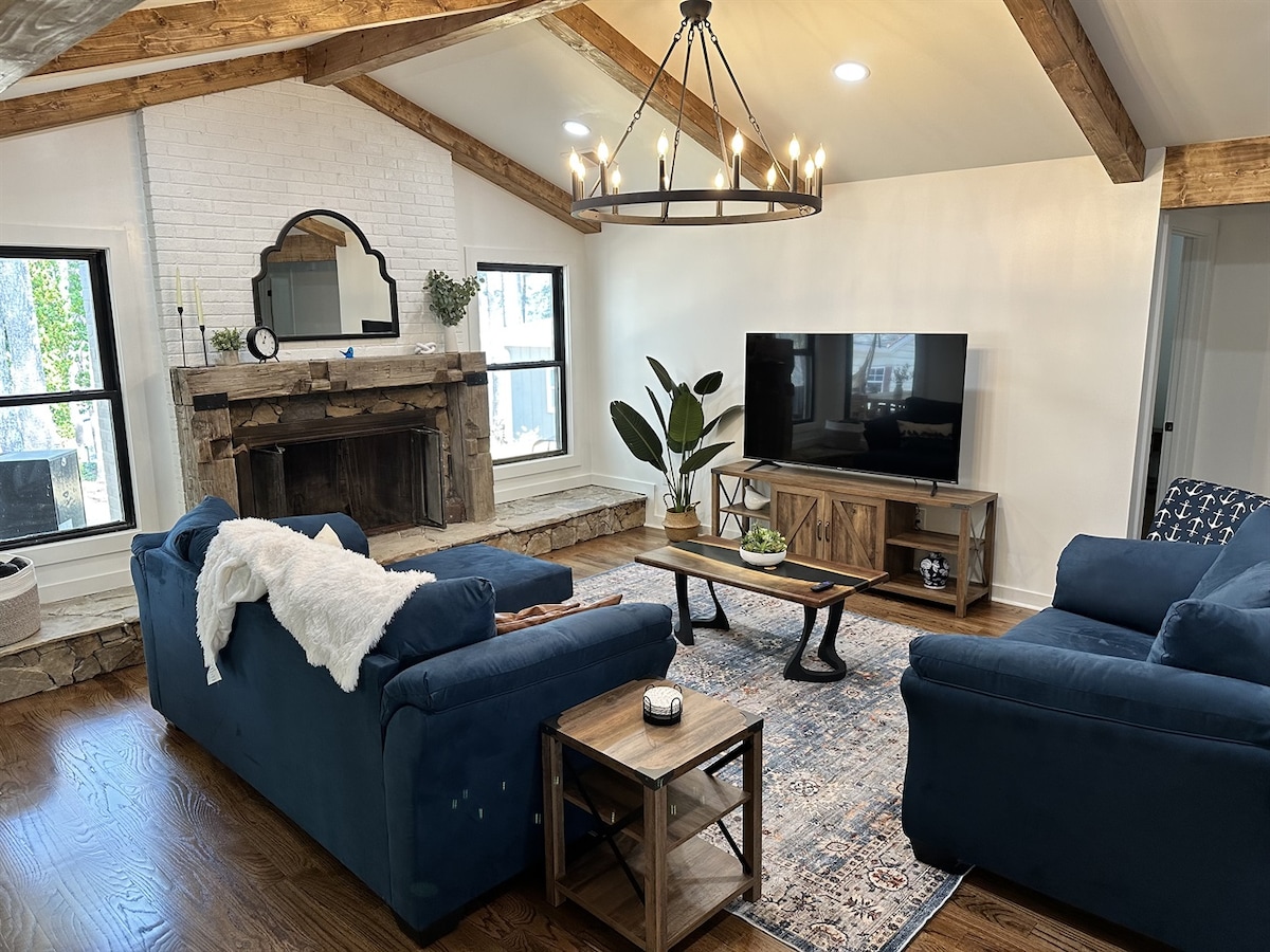 A family-friendly living space features two blue sofas positioned around a rustic coffee table. A stone fireplace with a wooden mantel serves as a focal point, complemented by a large television mounted on the wall. Natural light streams through the windows, illuminating the wooden beams above.