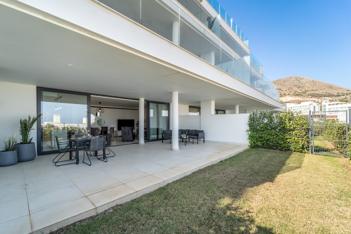 The outdoor terrace features a combination of seating and dining areas, providing ample space for gatherings. The well-maintained private garden features a lawn and is bordered by greenery, while large glass sliding doors connect to the living area, allowing natural light to fill the space.