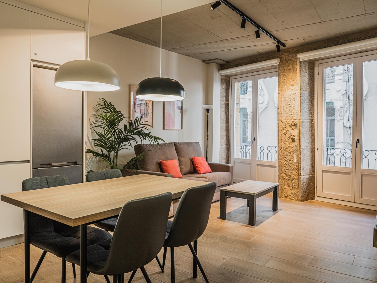A bright living area is featured, showcasing a wooden dining table surrounded by upholstered chairs. A cozy sofa with red accents is positioned against a wall, while large windows allow natural light to fill the space. Decorative plants add a touch of greenery.