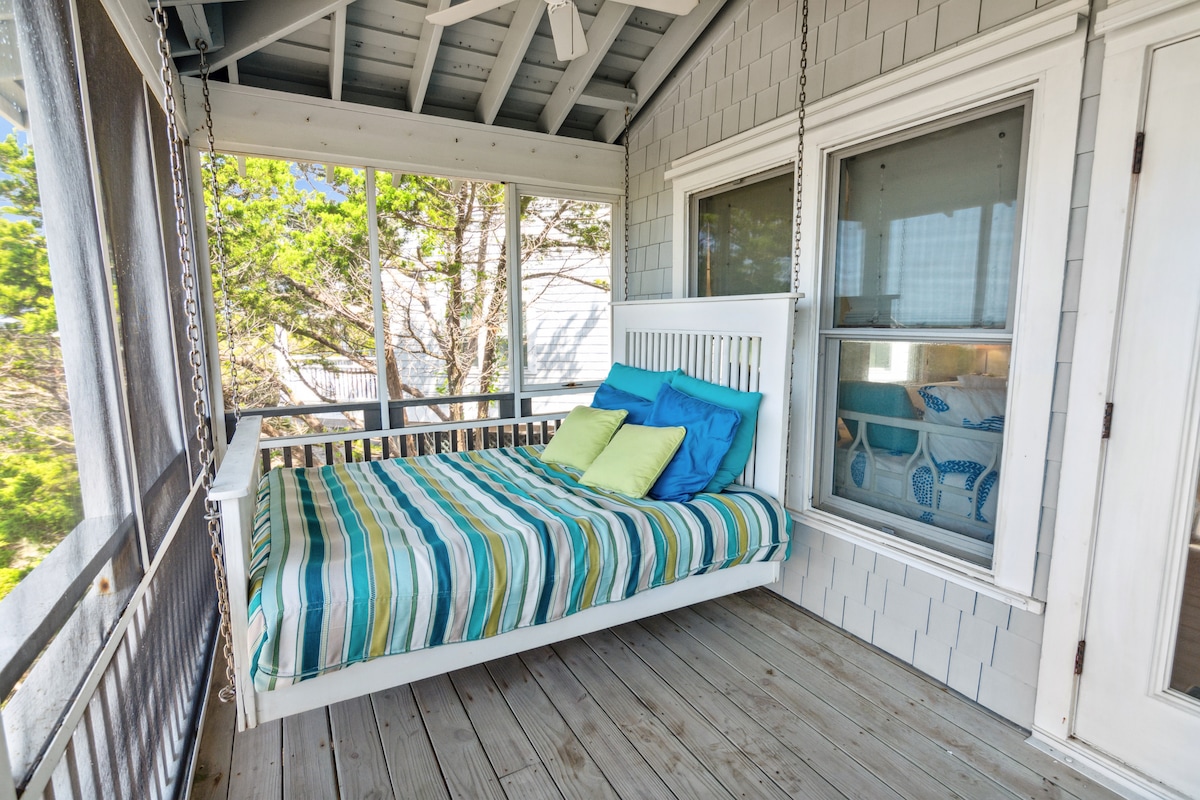 A cozy hanging bed is positioned on a screened porch, featuring striped bedding in shades of blue and green. Sunlight filters through the nearby windows, enhancing the tranquil setting. The surrounding trees provide a sense of being tucked away in nature.