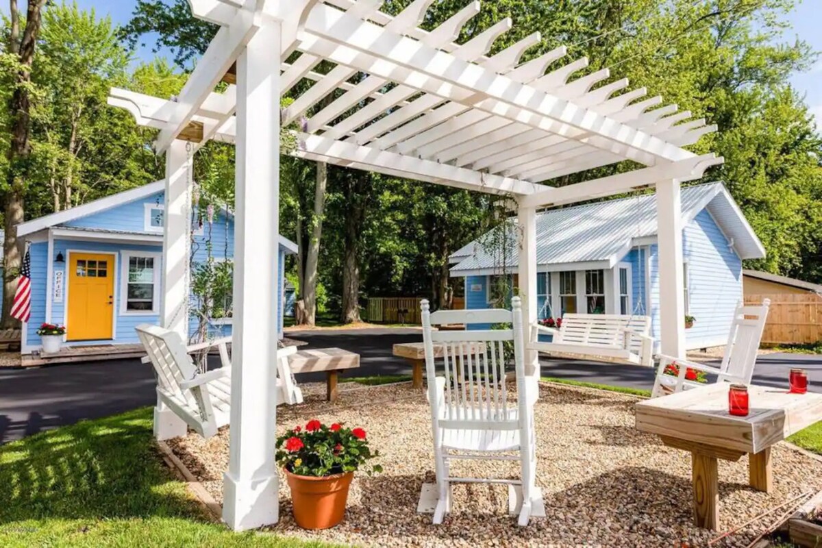 A white pergola provides shade over a gravel area, featuring a circular arrangement of four white rocking chairs. A round table, set with red candles, sits in the center. Potted flowers add a touch of colour to the surroundings, with blue cottages visible in the background.