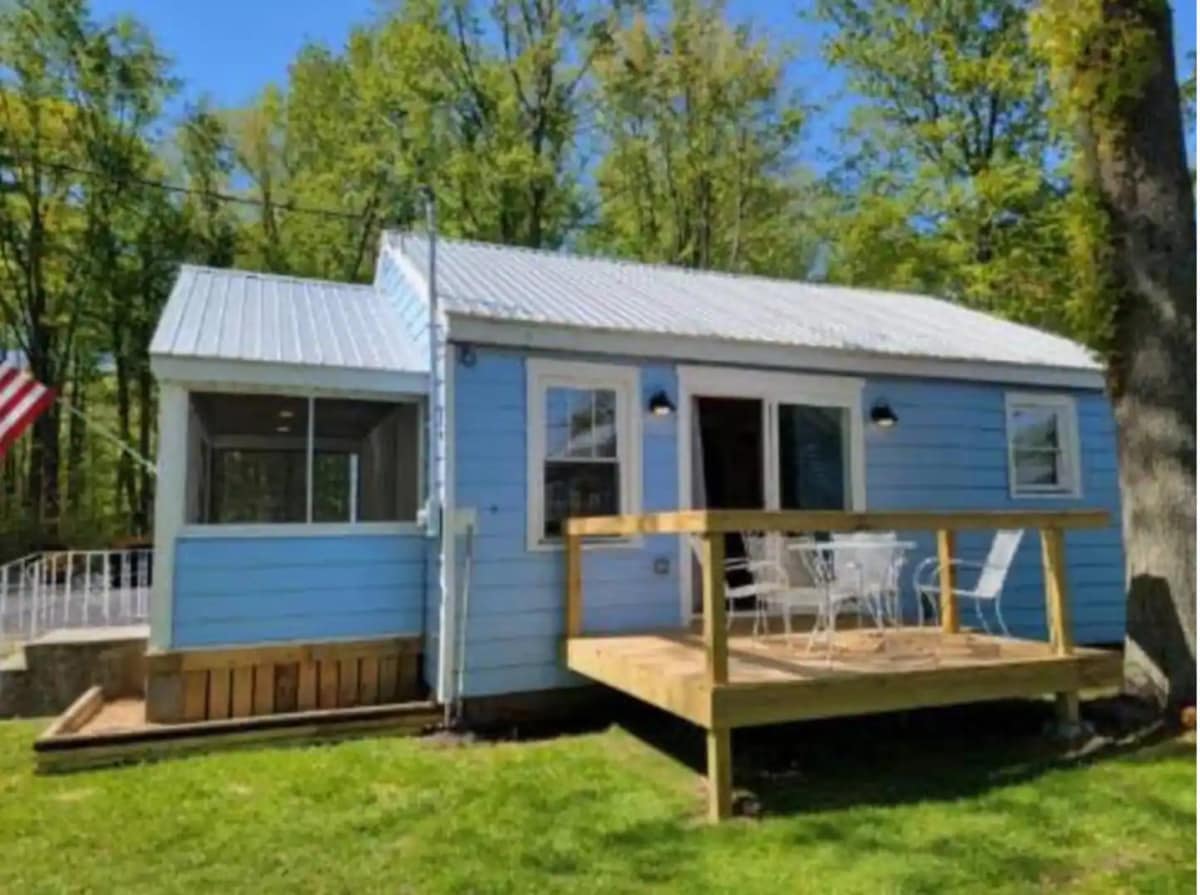 A charming cottage with a light blue exterior is set among greenery, featuring a wooden deck with outdoor seating. An American flag adds a welcoming touch, while large windows invite natural light into the space.
