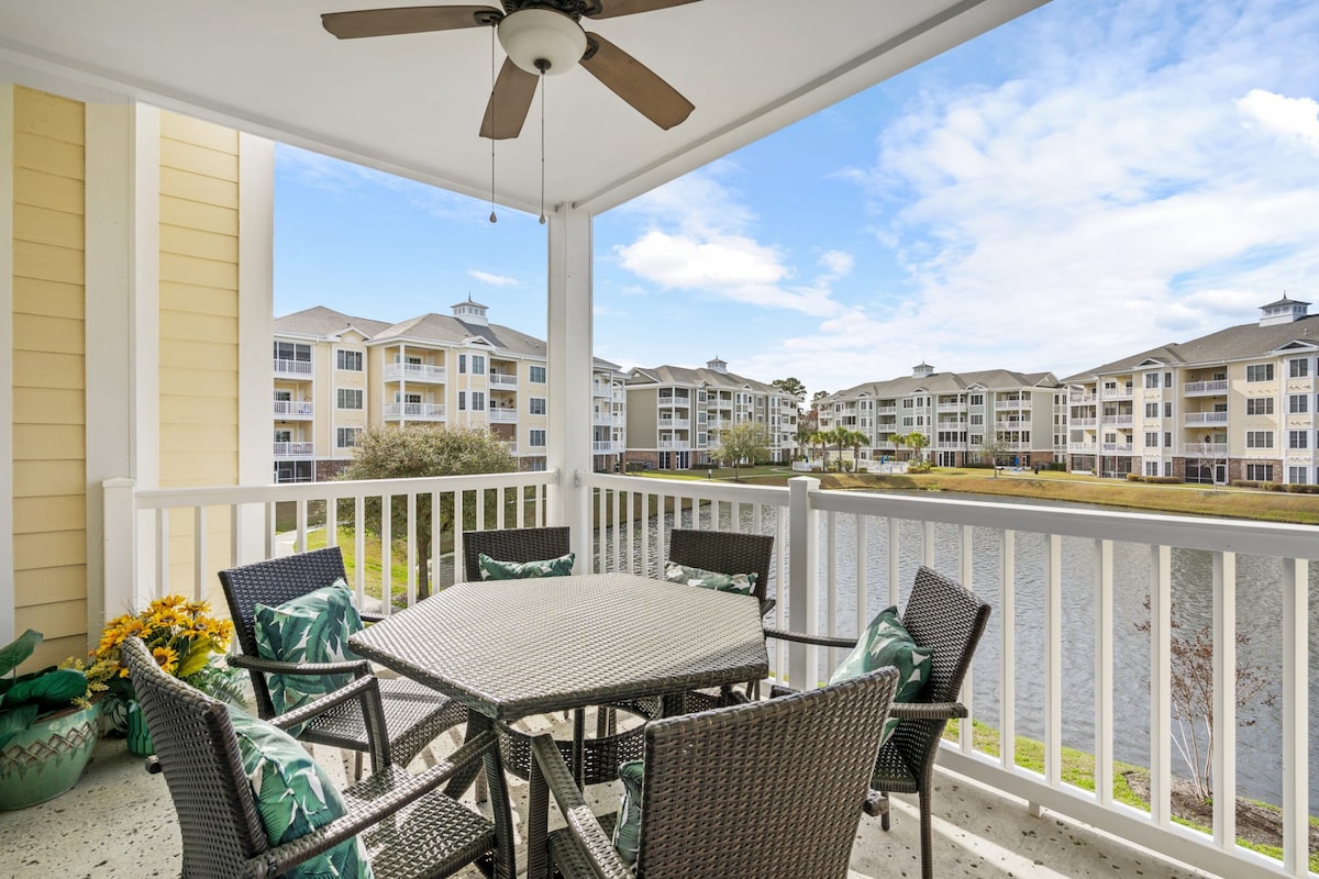 An outdoor balcony features a round dining table surrounded by six chairs, adorned with green and white cushions. The space overlooks a serene pond, with residential buildings visible in the background under a clear sky.