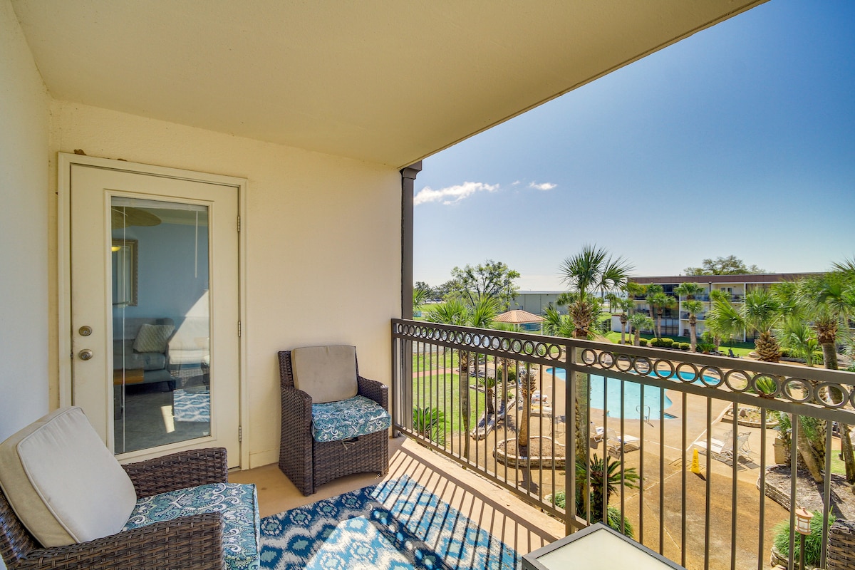 A private balcony features two cushioned chairs in a neutral tone, overlooking a landscaped area with palm trees and an outdoor pool. Natural light fills the space, highlighting the inviting outdoor seating arrangement and the clear blue sky above.