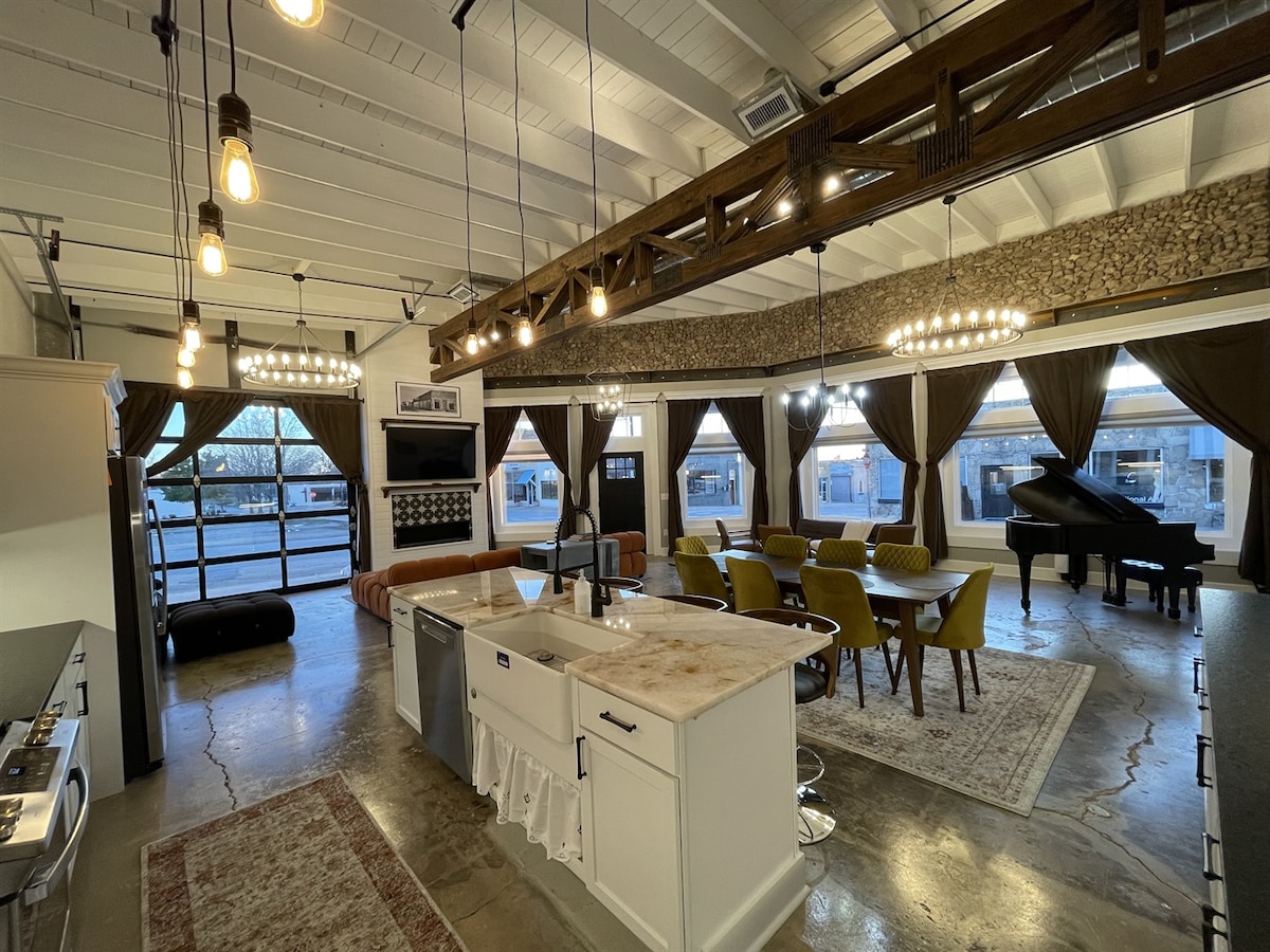 An open-plan living area features a modern kitchen with stainless steel appliances and an island. Dining tables with cushioned chairs surround the space, complemented by large windows dressed in elegant drapes. A grand piano adds a touch of sophistication beneath hanging light fixtures.