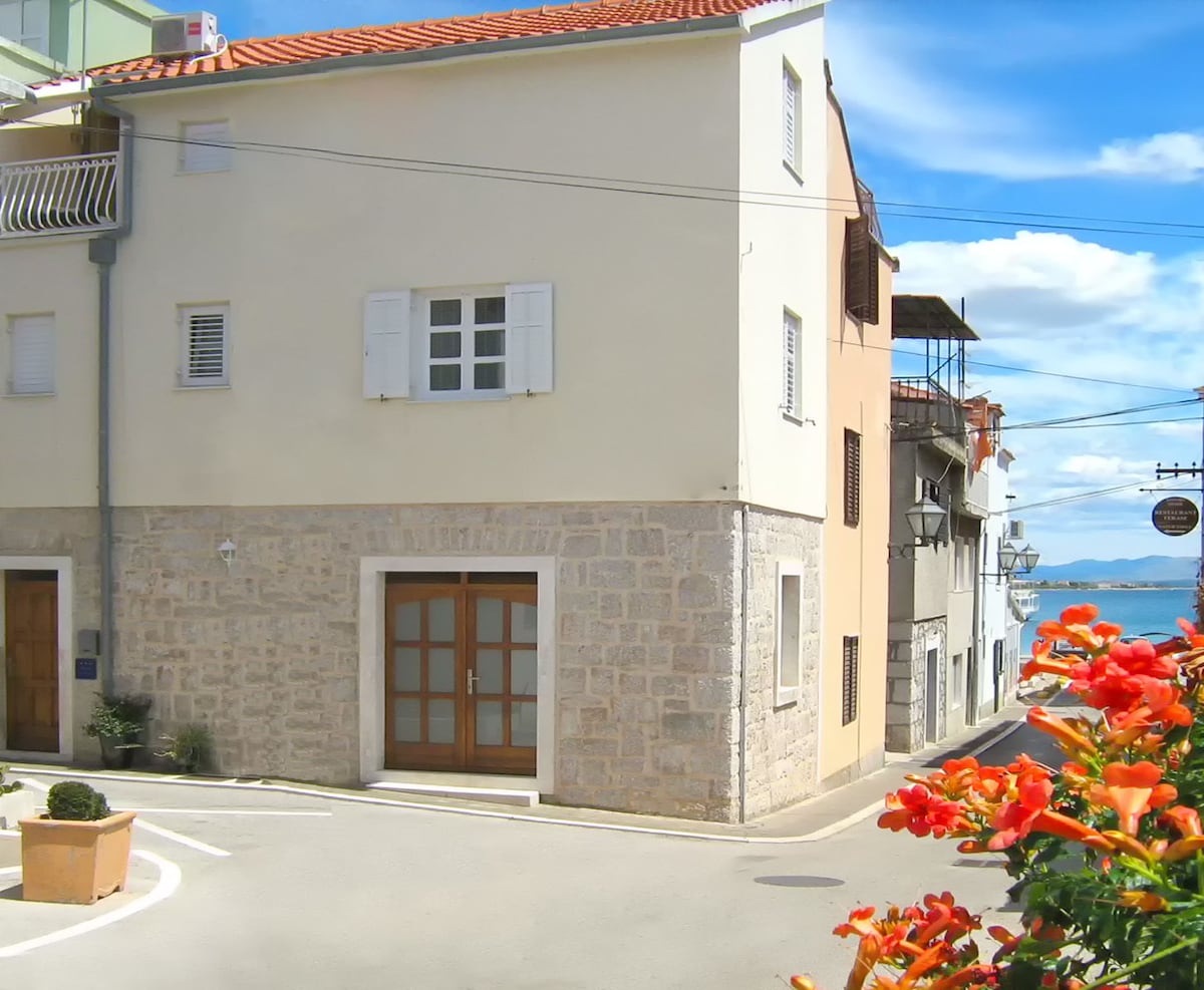 A corner building with a white façade and wooden window shutters is depicted. The ground level features a wooden door framed by stone, while vibrant flowers in a planter enhance the street view. The clear sky and distant water can be seen in the background.