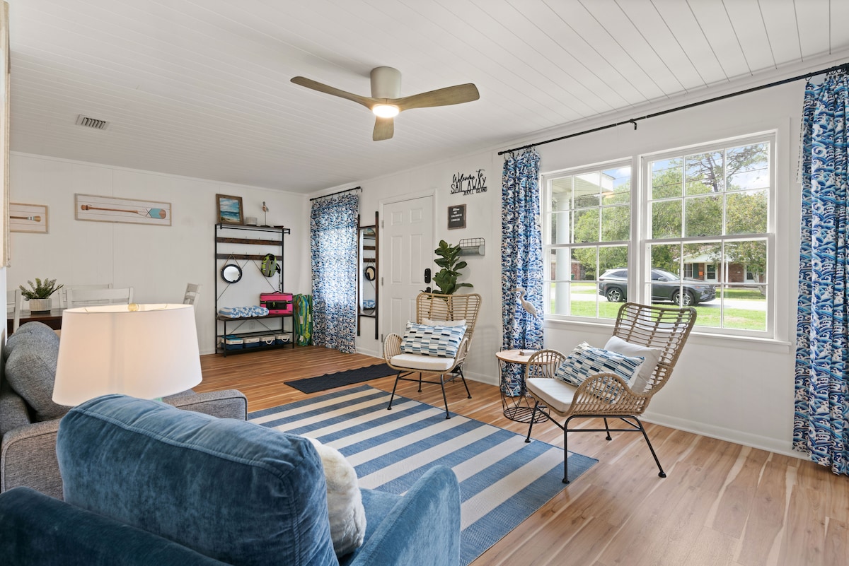 A spacious living area features a blue couch and comfortable rattan chairs on a striped rug. Large windows allow natural light to fill the room, with light blue curtains providing a pop of color. A ceiling fan adds functionality and comfort to the inviting space.