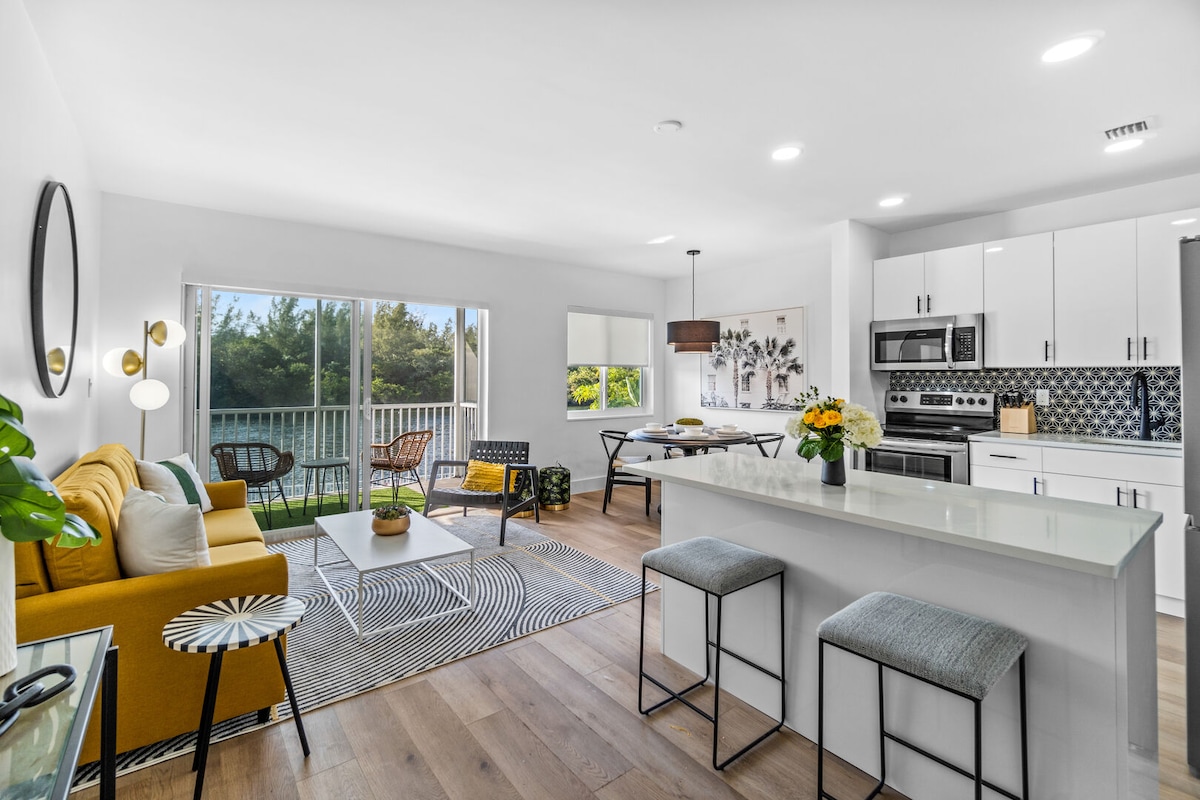 A stylish living area showcases a modern yellow sofa and patterned area rug, allowing ample natural light from large windows. A dining table complements the open kitchen equipped with sleek appliances. Decorative plants and artwork enhance the inviting ambiance of the space.