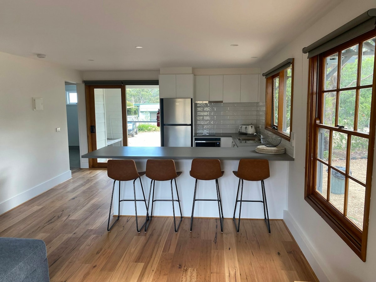 A modern kitchenette is showcased, featuring sleek cabinetry and a minimalist countertop. Four barstools are positioned at the counter, inviting casual seating. Large windows allow natural light to fill the space, while views of the outdoor area are visible through open glass doors.