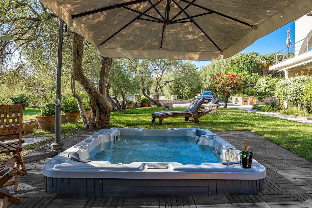 A shared jacuzzi is positioned under a large shaded umbrella, surrounded by lush greenery and outdoor lounge areas. Nearby, a wooden chair offers a place to relax, while blooming flowers add natural beauty to the garden setting.