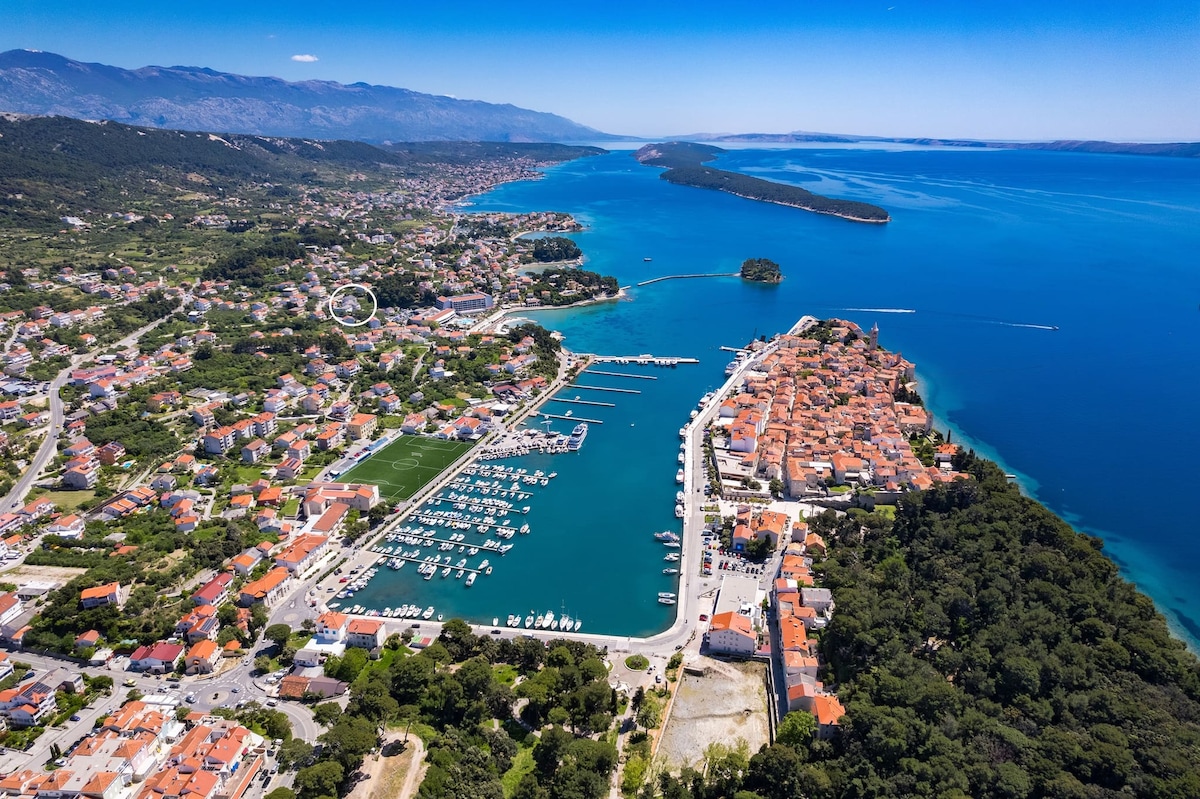 An aerial view captures the vibrant coastline of Rab island, featuring a mix of green hills and residential areas. The marina is visible with numerous boats docked, while the clear blue waters reflect the sunlight. The landscape seamlessly transitions from urban development to natural surroundings.
