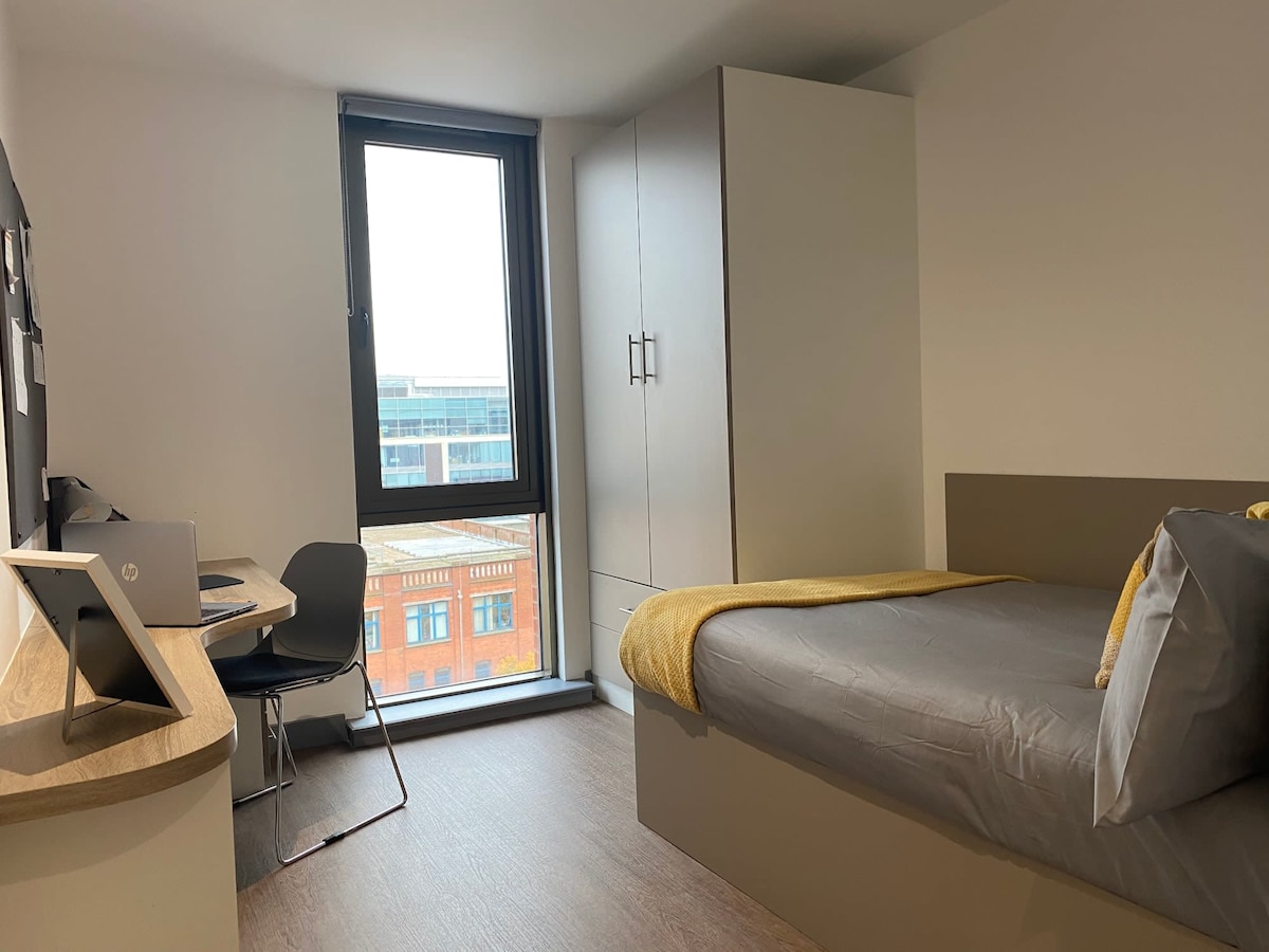 A modern studio features a spacious bed with a neatly arranged duvet, complemented by a vibrant yellow throw. A study area with a desk and chair is positioned near a large window, allowing natural light to illuminate the space. Ample storage is provided by a built-in wardrobe.