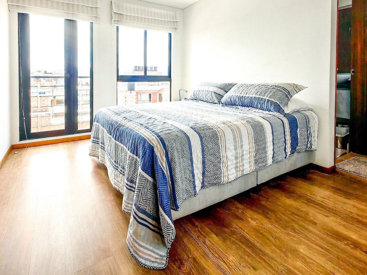 A well-lit bedroom features a spacious bed with an intricately patterned blue and white blanket. Large windows provide natural light and offer views of the surroundings. The modern flooring complements the neutral wall color, creating a bright and open atmosphere.